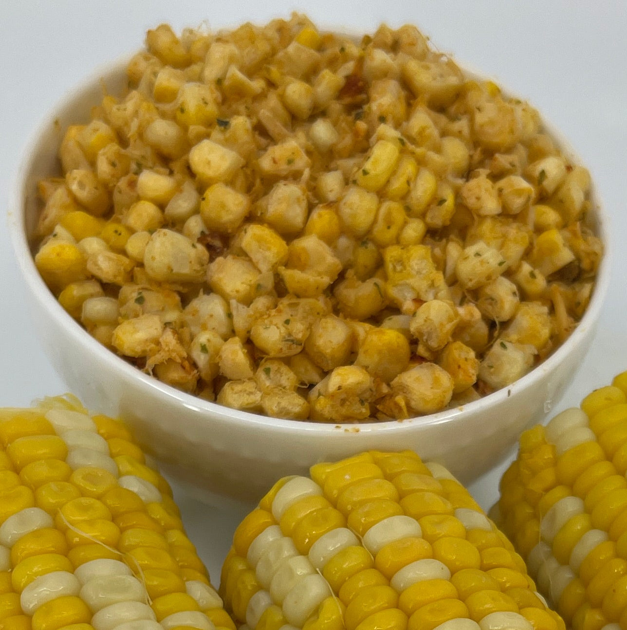 Three ears of corn on a white plate with a small bowl of yellow kernels on a white background